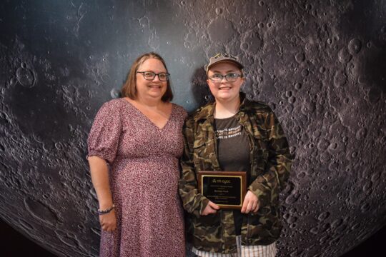 A mom in a dress stands next to her daughter in a camouflage cap and shirt holding an award plaque