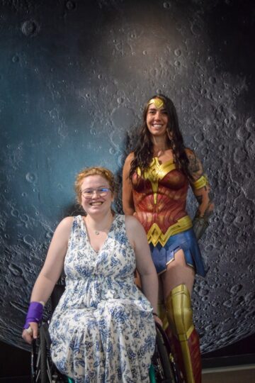 a teenage girl in a long white and blue paisley dress sitting in a wheelchair next to a standing Wonder Women in costume. 