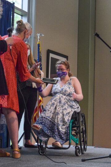 A teenage girl in a long blue and white paisley dress sitting in a wheelchair on a stage accepting an award plaque from a woman standing in a red dress 