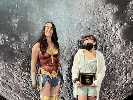 A tall woman with long dark hair is dressed as Wonder Woman standing next to a teenage girl holding an award plaque. 