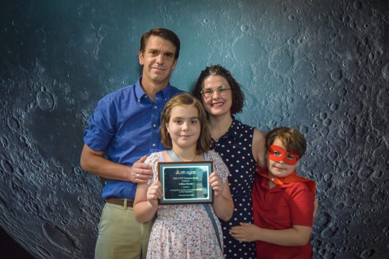 A family consisting of a Dad, Mom, Daughter and son standing together. Dad has his hand on his daughters shoulder and she is holding an Award Plaque. The son is wearing a superhero mask and cape.