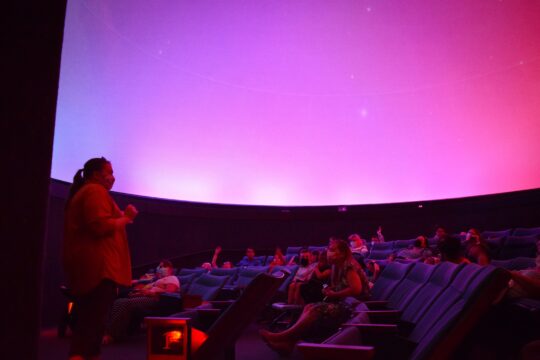 A purple darkend room with kids sitting in auditorium seating listening to a speaker up front 