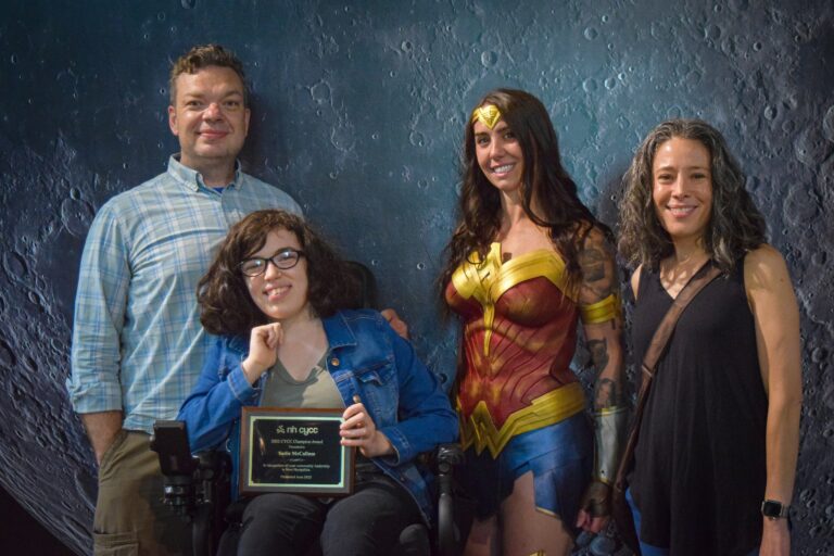 A dad standing behind his daugher in a wheelchair. She is holding an award plaque. Next to her is Wonder Woman and her mom, all smiling.