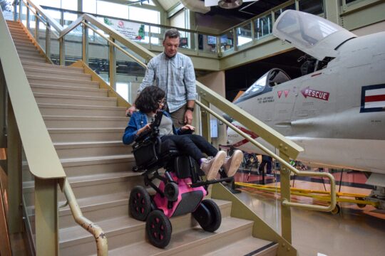 Teenager in a specialized pink wheelchair using the stairs with a man looking on behind her