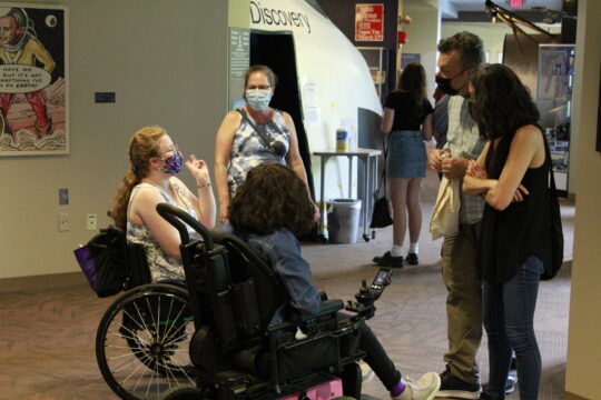 Two teen girls in wheelchairs talking with standing adults standing in a circle 