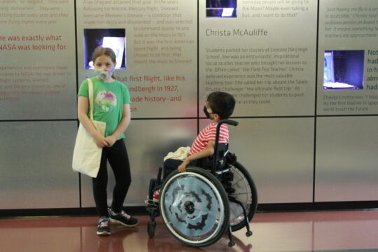 Child on left looking straight ahead with bag on shoulder wearing a mask standing next to young boy in wheelchair looking at wall exhibit