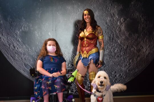 wonder woman standing next to a girl wearing a blue top and flower skirt in a wheelchair. The girl is holding a leash for a white curly dog. 