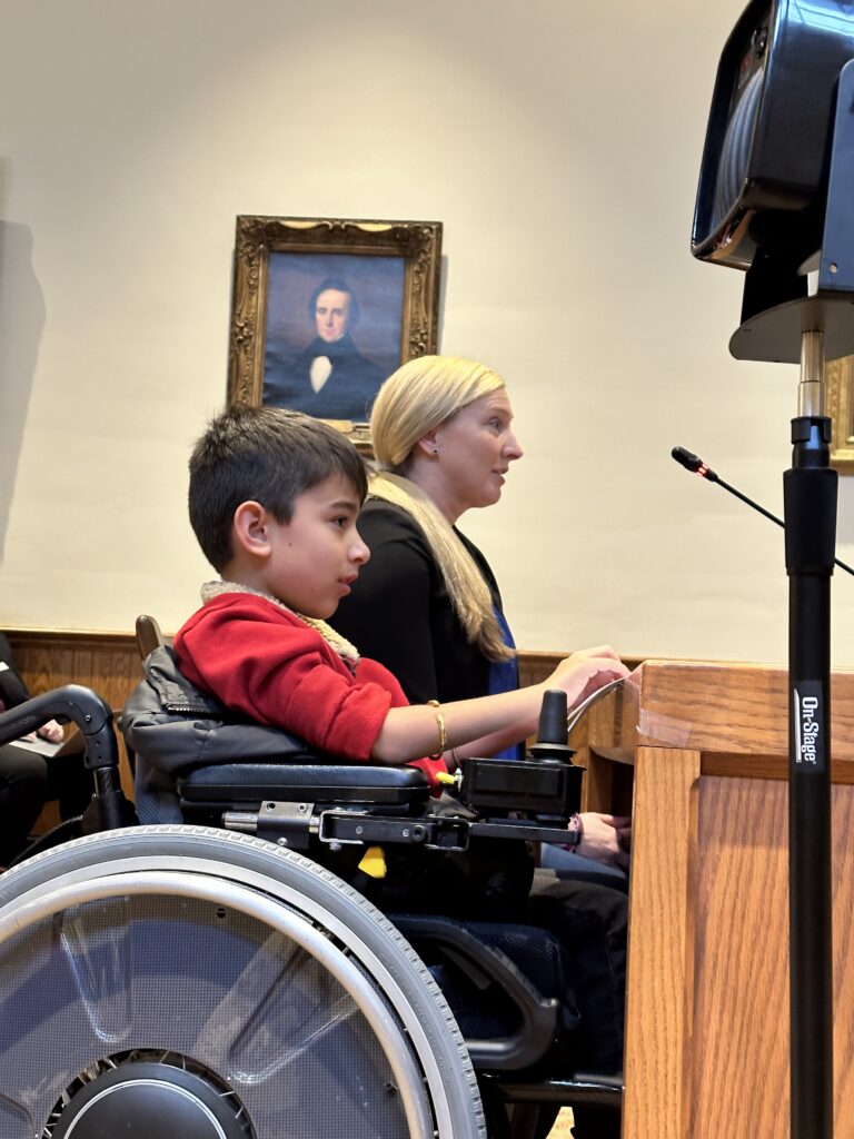 A young boy with short brown hair and a red tshirt sits in a wheelchair next to his mother with long blond hair and black clothing. Its a side view sitting at a desk with a microphone.