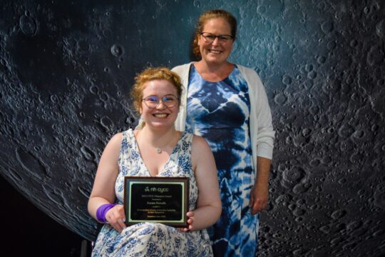 A teenager girl sits in a wheelchair wearing a blue flowered dress holding an award plaque. Her mom stands behind her wearing a blue tie-dye dress.
