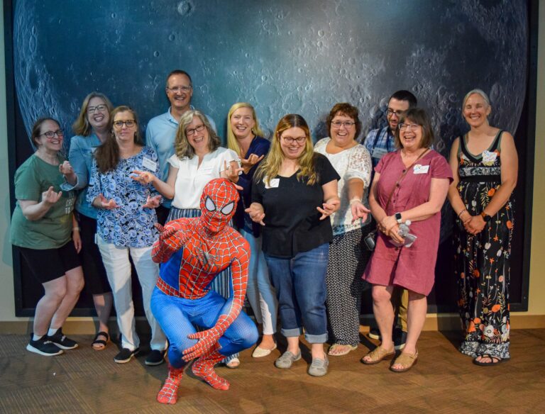 A group of adults laughing and reaching arms out to spin a web, while Spiderman sits posed in front of them